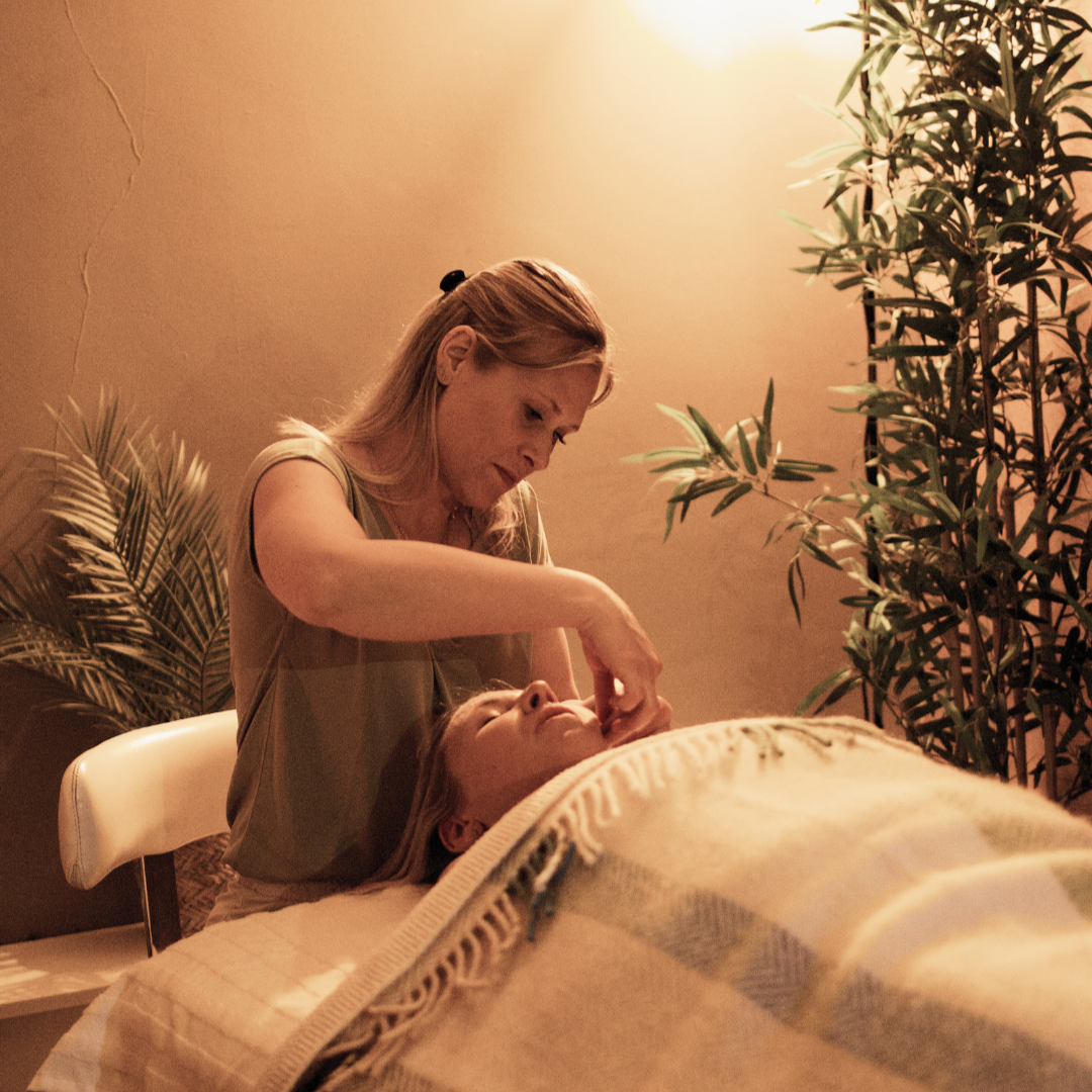 A person receiving a facial massage from a massage therapist in a dimly lit room with plants in the background.